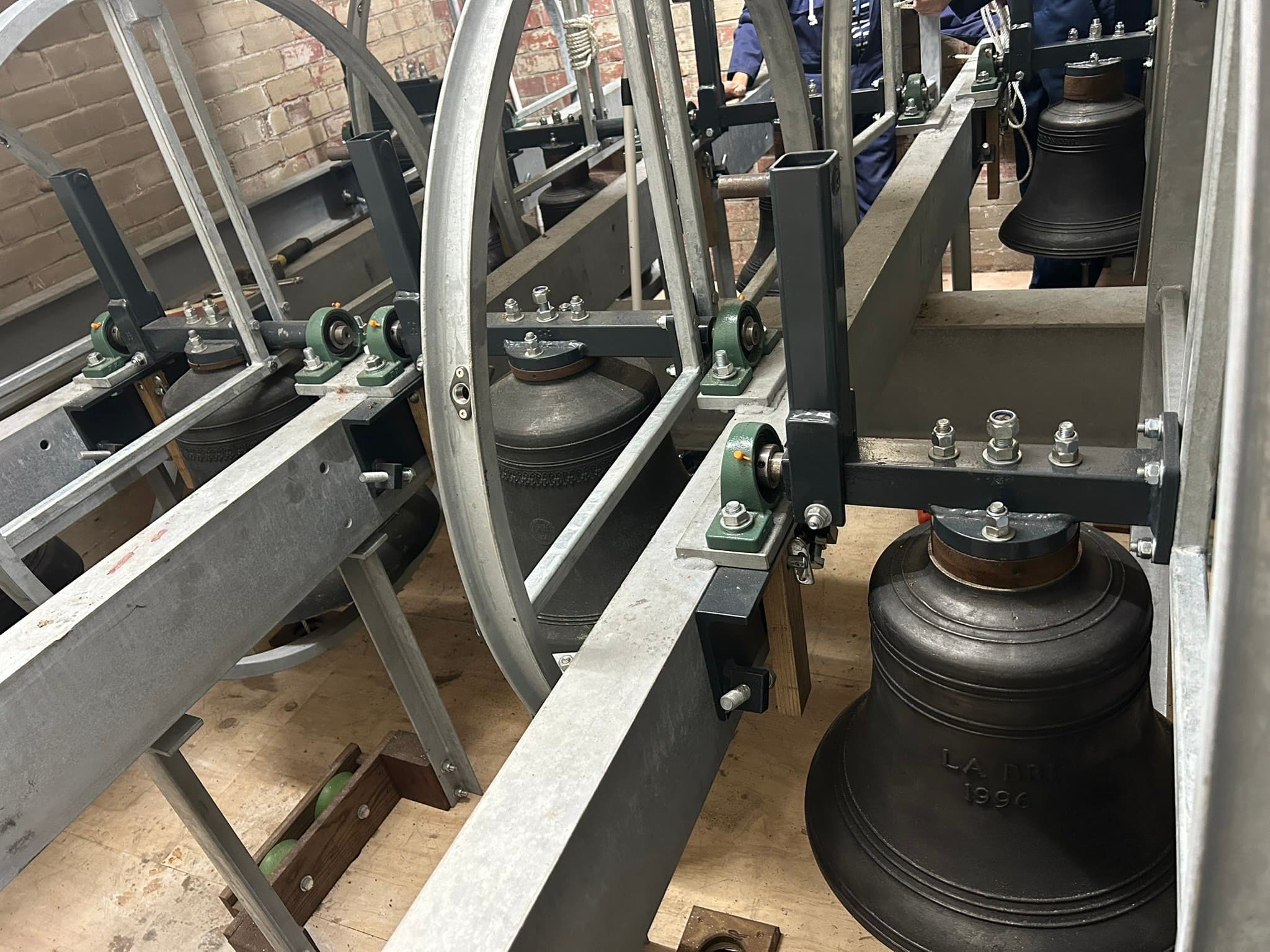 A new galvanised steel bell frame bolted into the brick wall of a church tower, bearing blocks and fixings visible.