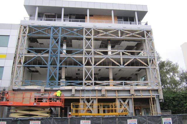 A multi-storey building stripped back to its floor slabs and columns, with a steel braced frame bolted across the full height of the elevation. A worker stands on a raised scissor lift at ground level, behind site fencing hung with hazard notices.