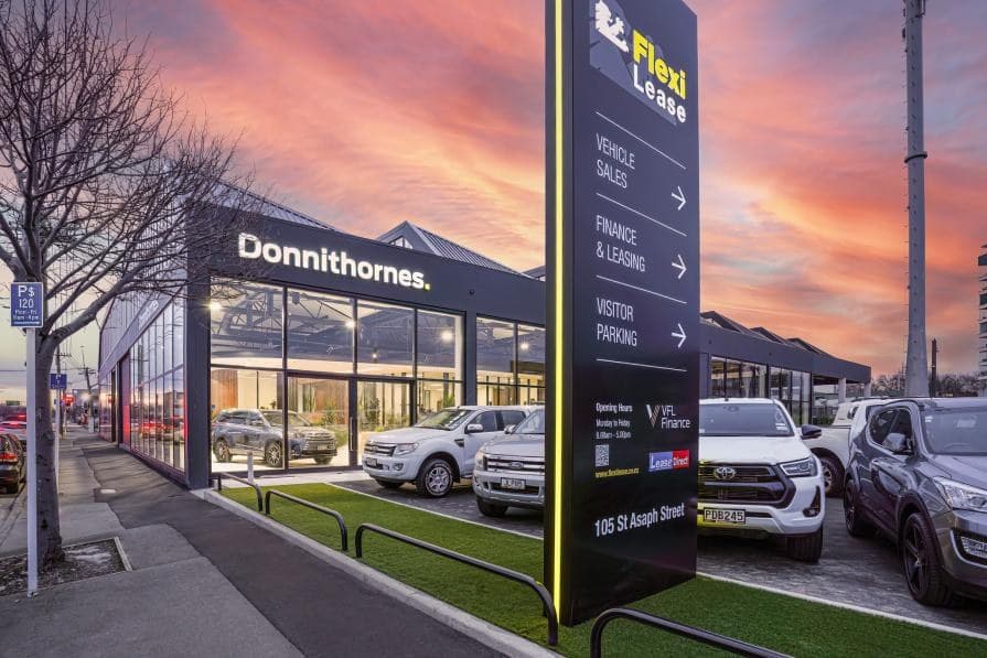 The building from the street at dusk, a lit dealership frontage behind a forecourt of vehicles, with a tall illuminated sign at the kerb.