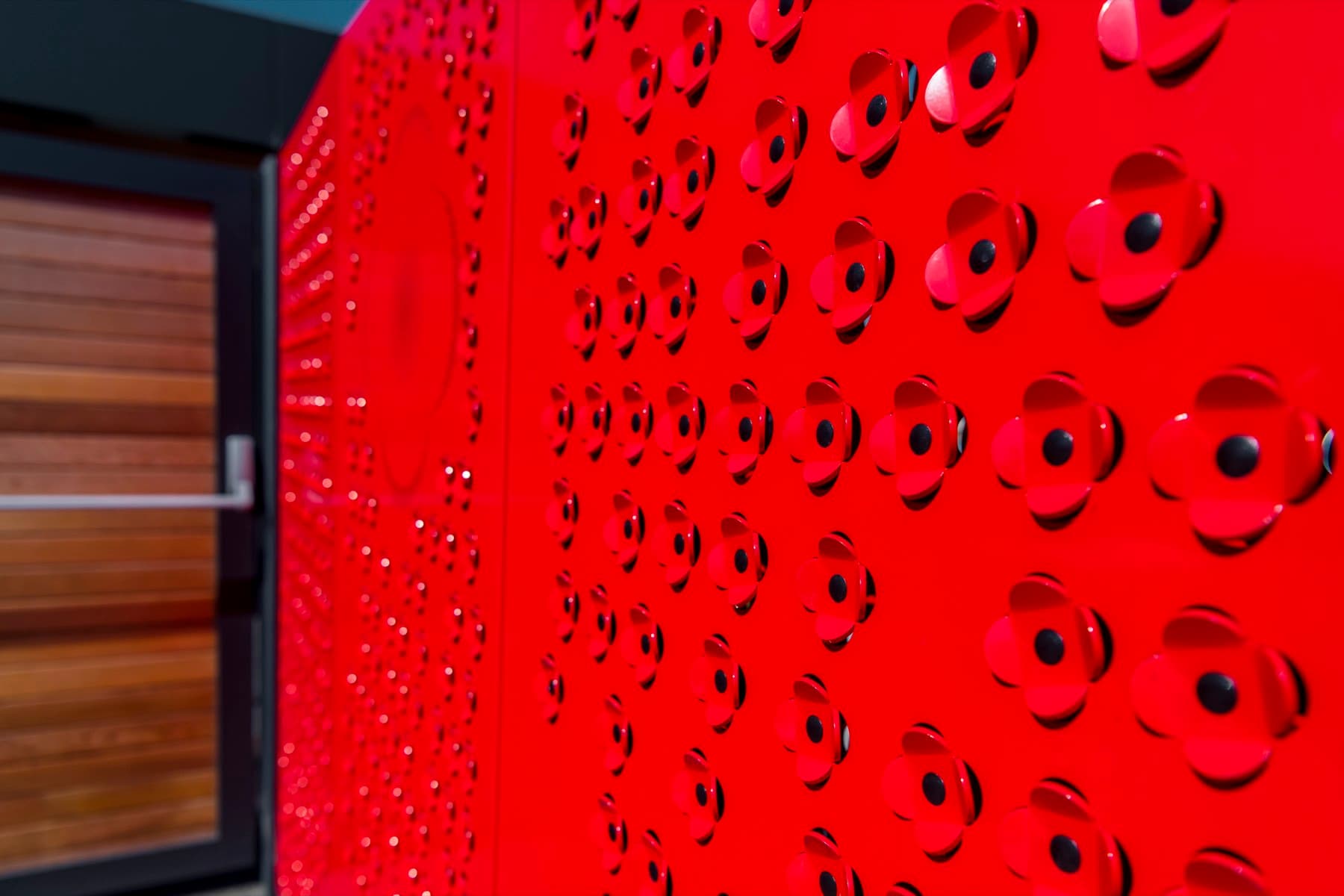 A red powder-coated screen formed and perforated as rows of poppies, set against timber cladding.
