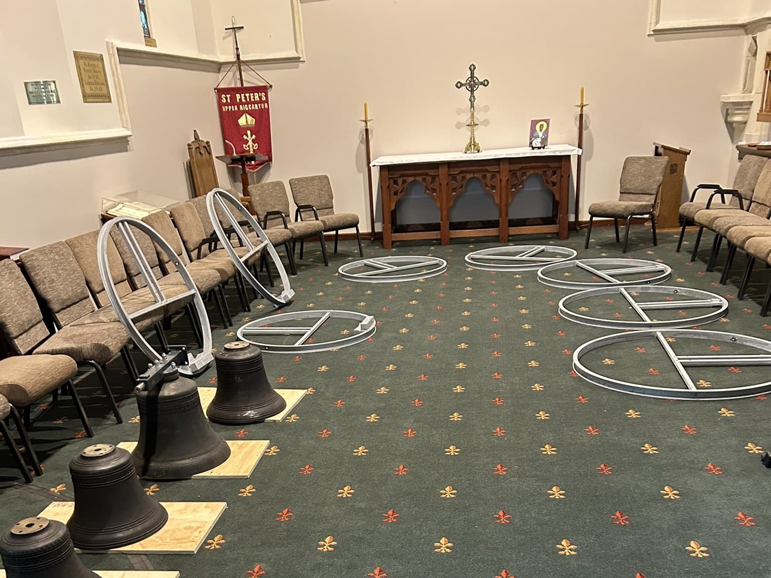 Four bronze bells standing on plywood pads on the patterned green carpet of a church interior, one of them still bolted to a black steel headstock, with circular galvanised steel wheel frames laid out flat across the floor around them, stacking chairs pushed back to the walls, and an altar table with a brass standing cross and candles beyond