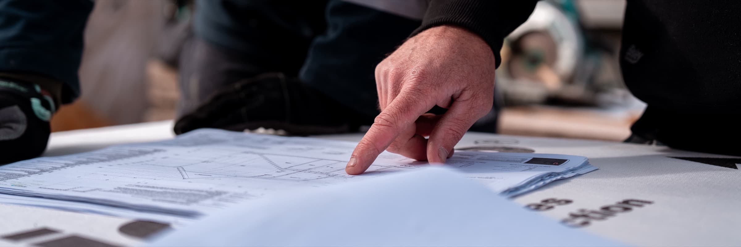 A hand pointing at a set of building drawings spread across a table, a hi-vis sleeve above them and a second person standing at the edge of the frame