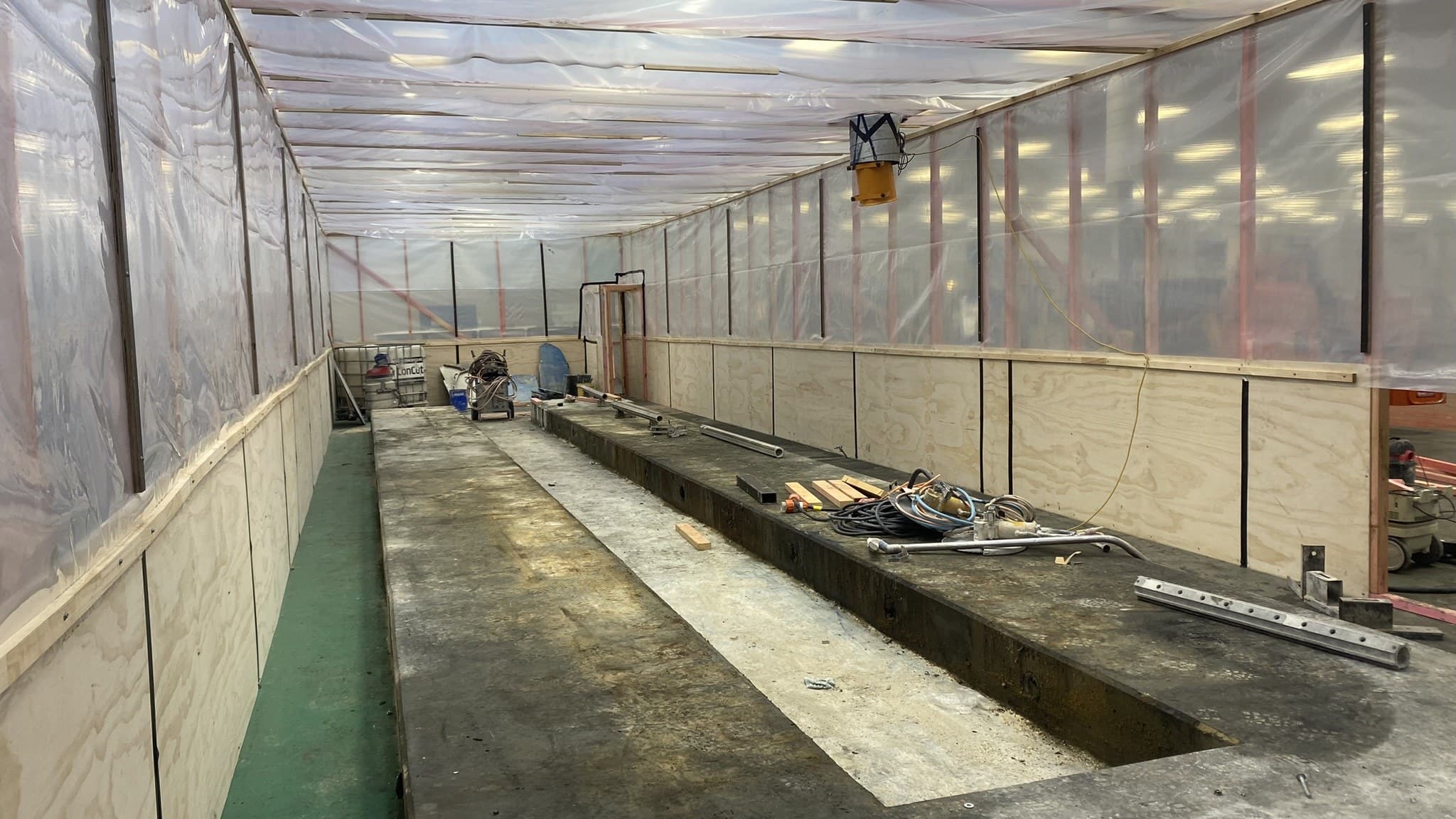 A timber-framed plywood and polythene dust screen dividing a work area from the rest of an operating food-processing plant, the plant's lit floor and racking visible through the screen