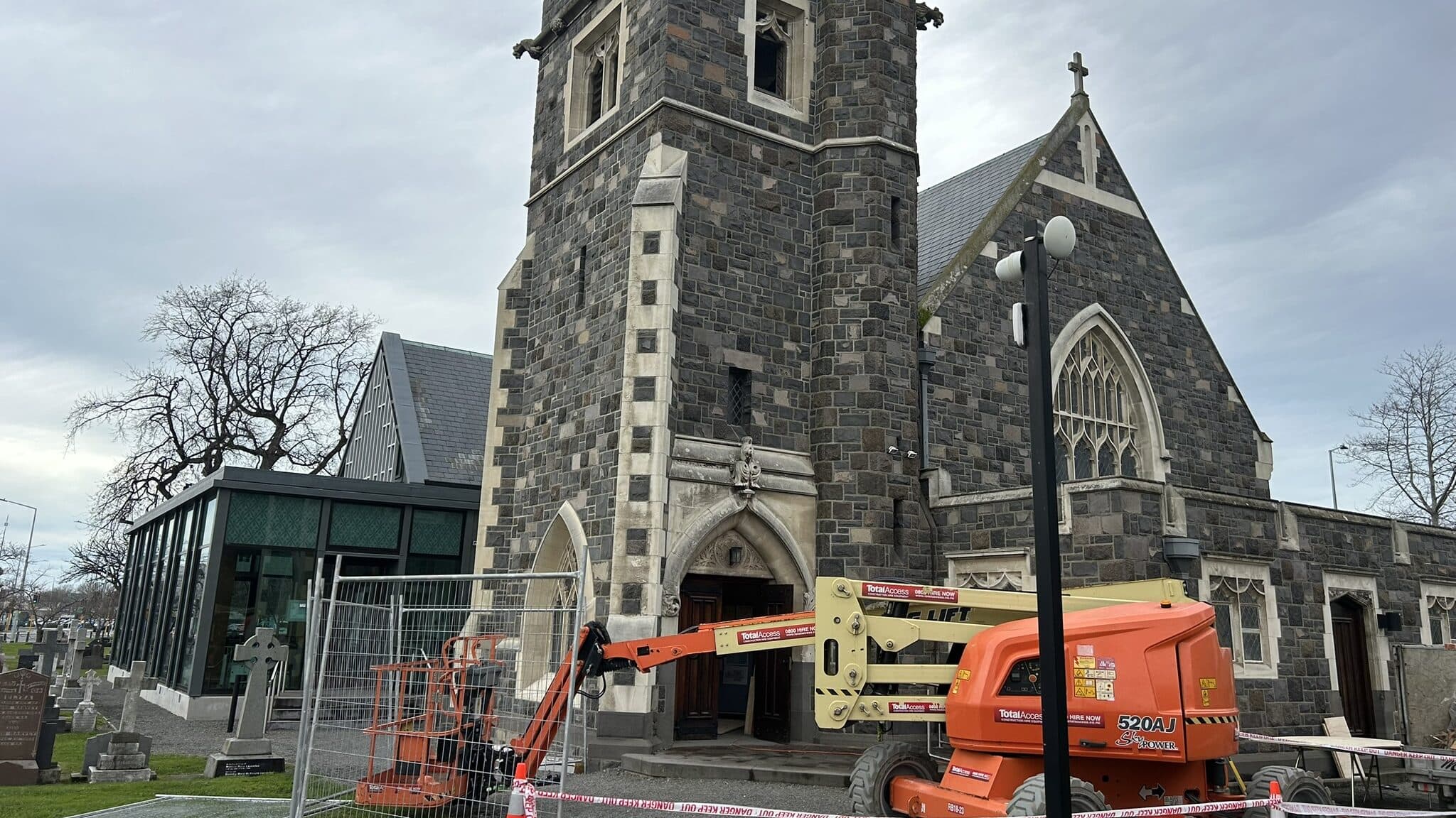 Temporary mesh fence panels and DANGER KEEP OUT tape closing off the frontage of a stone church, a boom lift standing at the open entrance doors