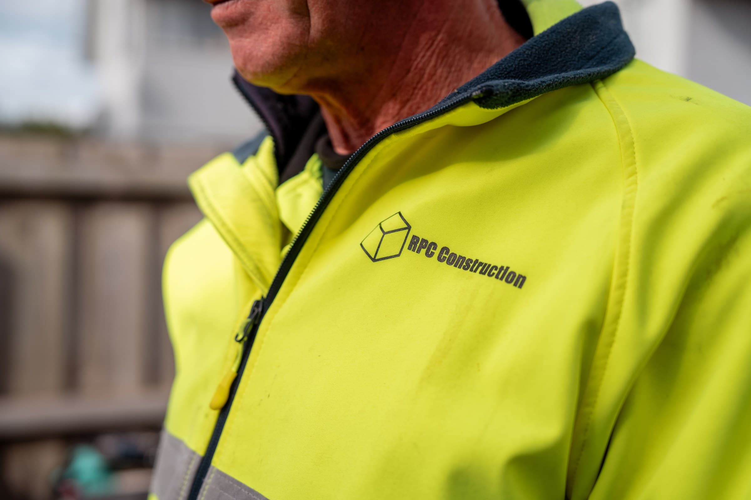 A close view of the shoulder and chest of a high-visibility yellow work jacket worn on site, with the RPC Construction cube logo and wordmark printed on the chest, the wearer's face out of frame above