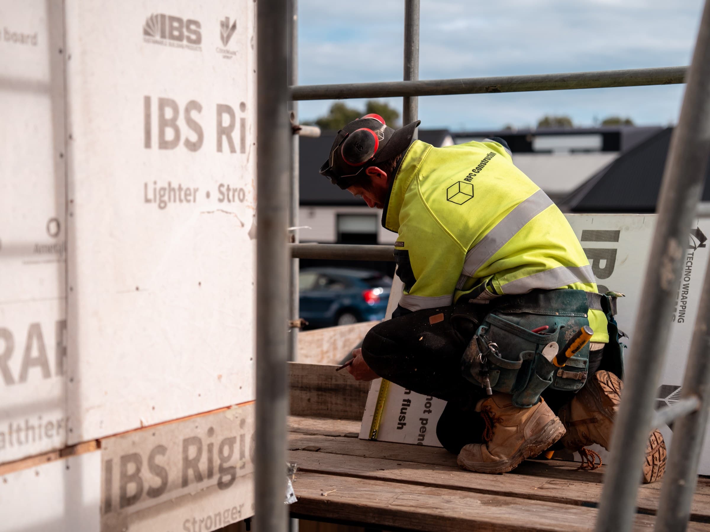 A worker in RPC Construction hi-vis and ear defenders kneeling on a scaffold plank, marking a line with a steel tape and pencil, sheets of rigid wall board stacked beside him and scaffold rails across the frame