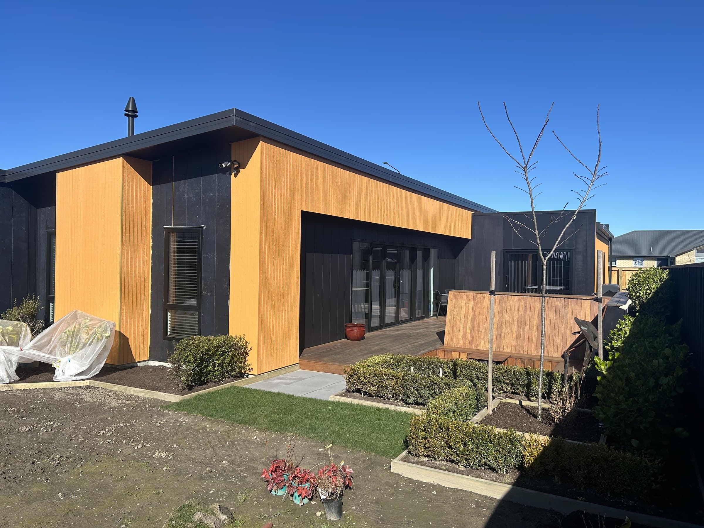 A finished single-storey house with dark board cladding and vertical cedar panelling, a timber deck along the front and clipped hedging beside a paved path