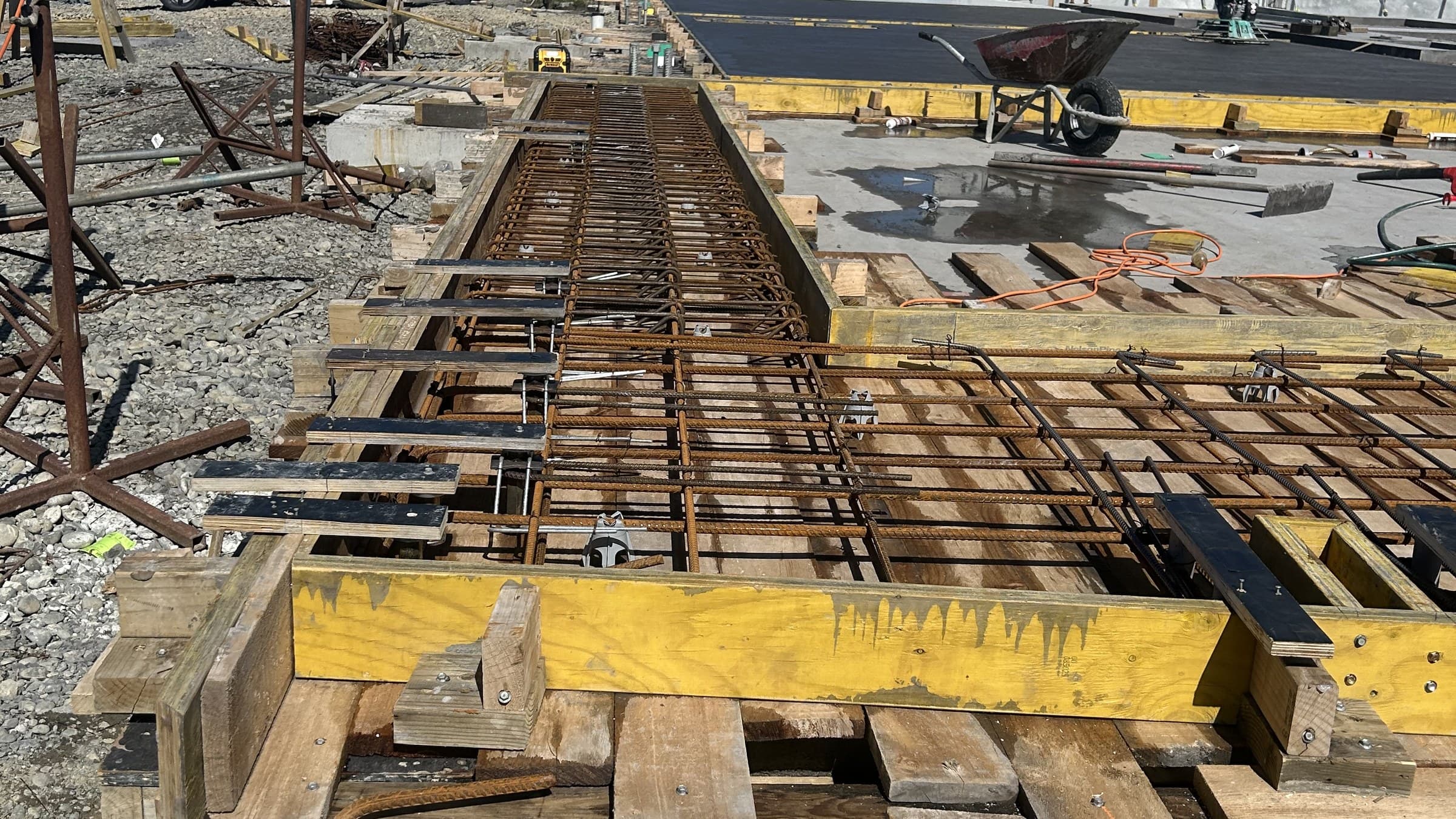 Reinforcing steel tied into timber formwork for a long foundation beam on an open site, with precast concrete panels standing beyond and a wheelbarrow on the poured slab