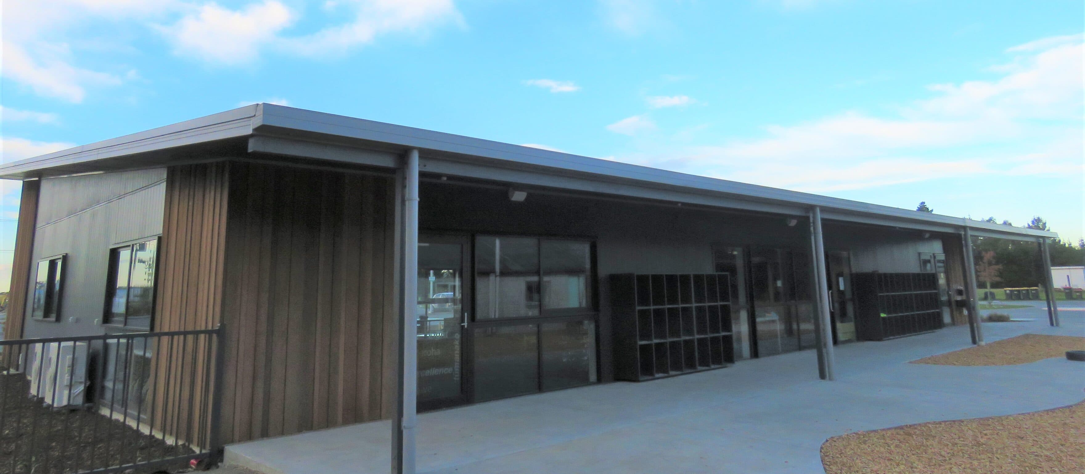 The completed cedar-clad teaching block at Rolleston Christian School in daylight, a covered walkway on slender steel posts running the length of its front, a bank of empty cubby shelving standing against the wall, the glazed doors closed and the concrete path empty