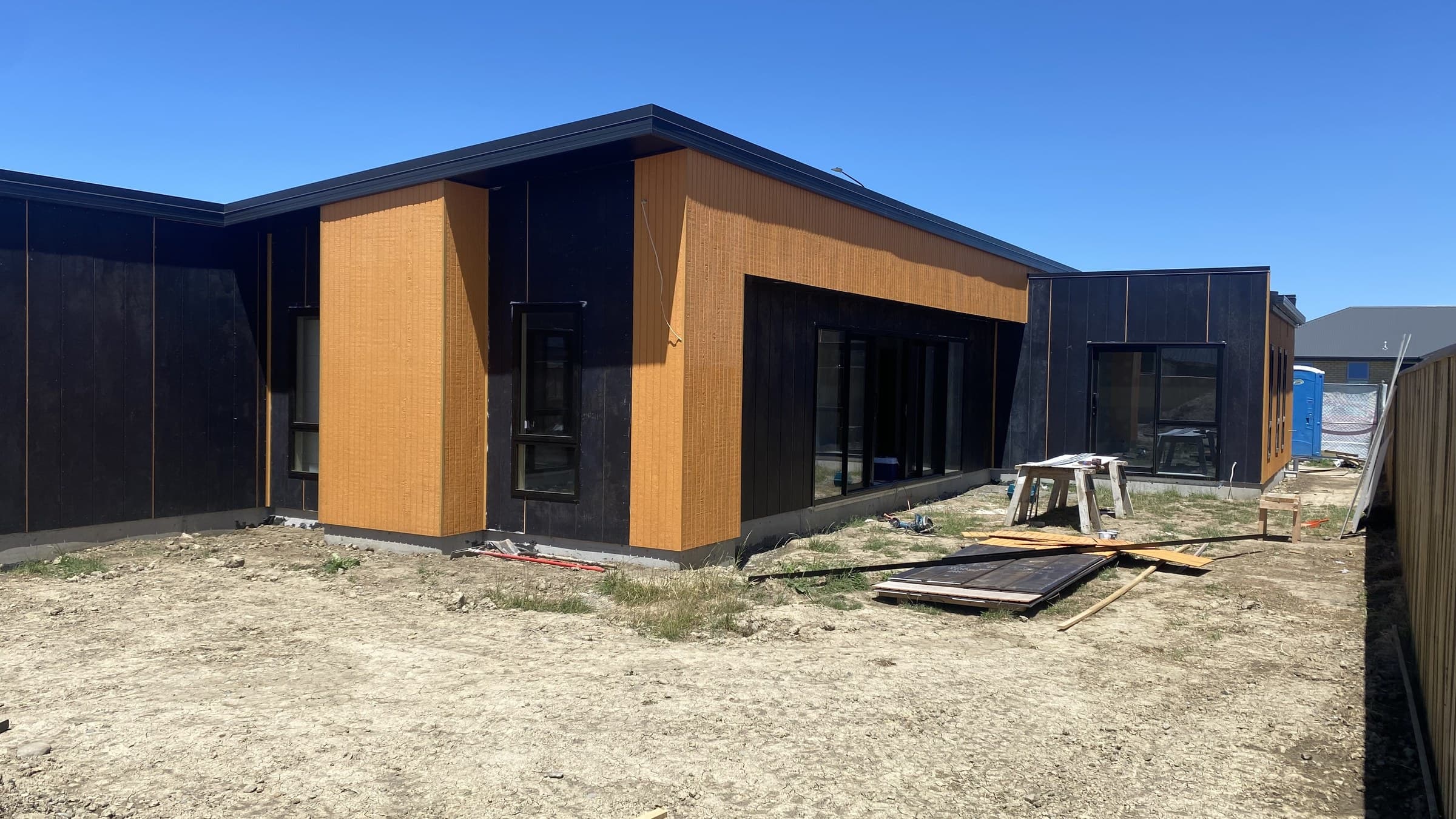 A newly completed single-storey teaching block clad in dark board and vertical cedar panels, standing on bare unlandscaped ground with trestles and timber offcuts still lying about