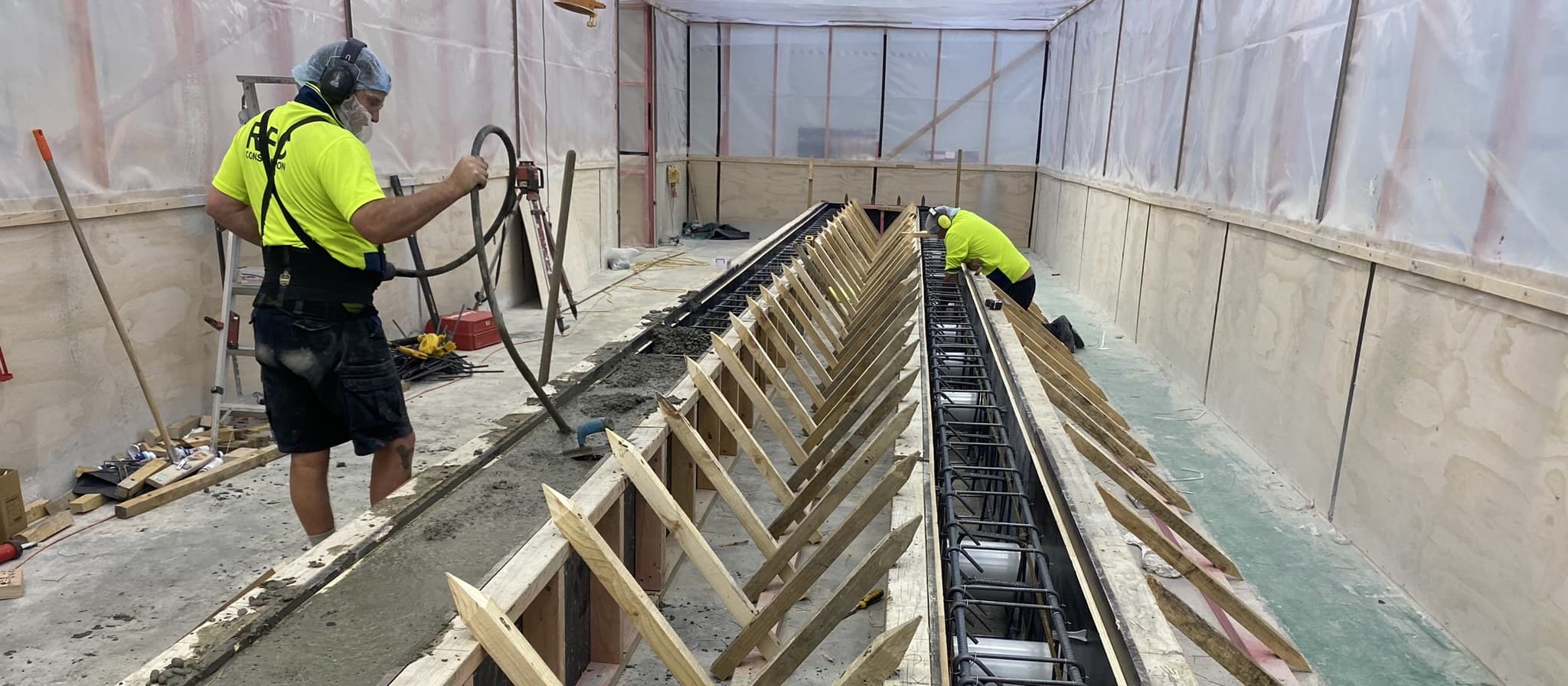 Two workers in RPC Construction hi-vis, hairnets and dust masks forming a long in-situ concrete channel, timber formwork and angled props running its length, a reinforcing cage in the open trench, and the work area sealed off behind plywood and clear sheeting