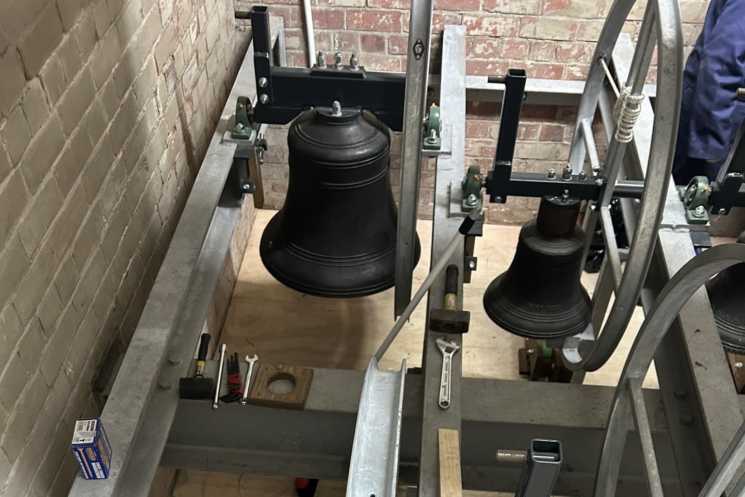 An installed galvanised steel bell frame in a brick belfry, bells hung on their headstocks with bolted fixings and green bearing blocks, a wheel alongside and hand tools laid along a girder