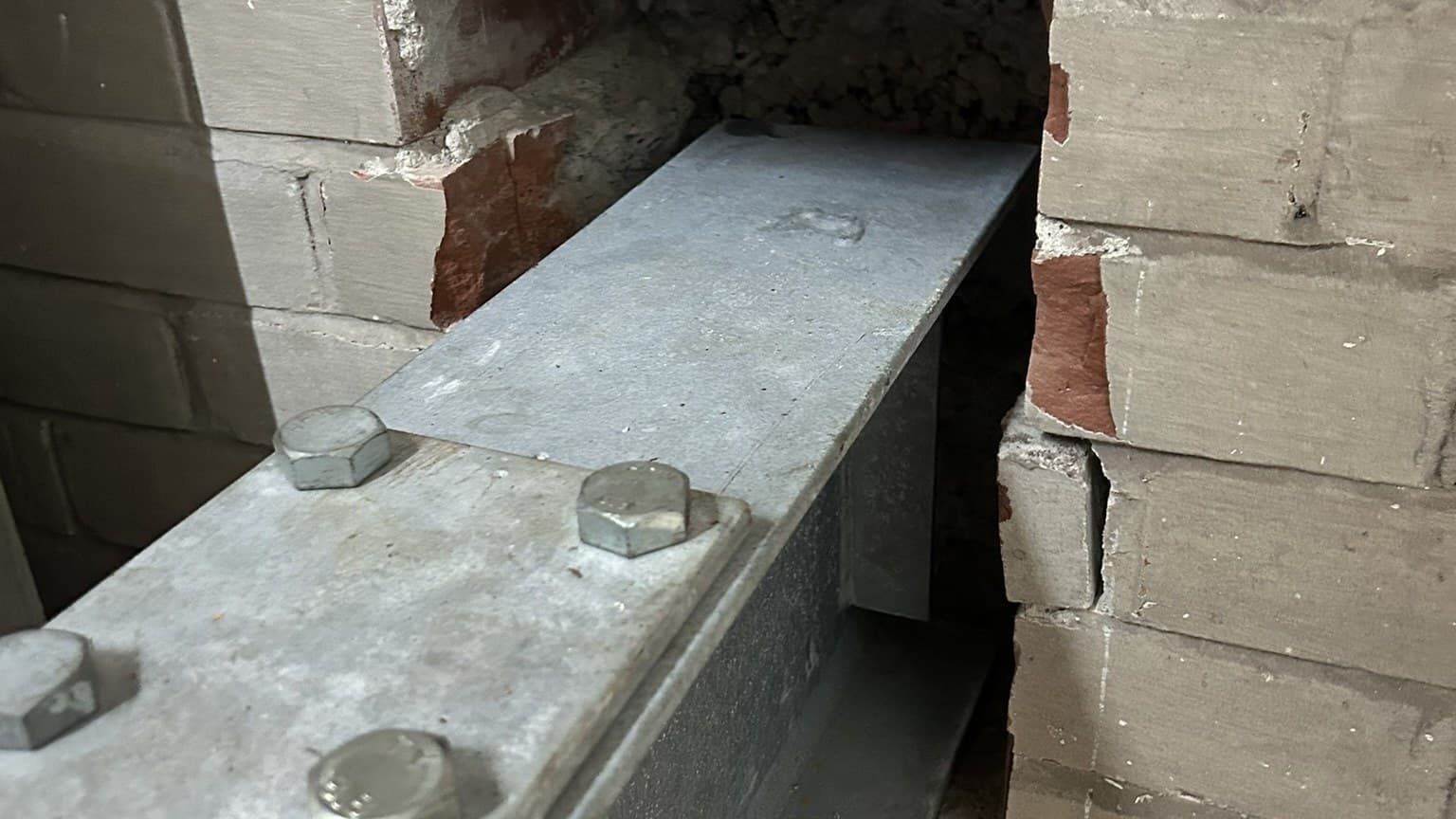 A galvanised steel beam bolted through a plate into a brick wall, with the brickwork cut away around the connection and older red brick exposed behind it