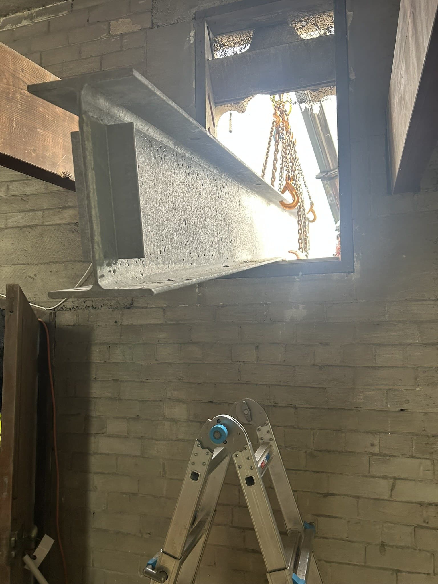 A galvanised steel beam being fed through an opening cut high in a whitewashed brick wall, lifting chains and hooks hanging in the daylight beyond it, with a folding ladder standing against the wall below