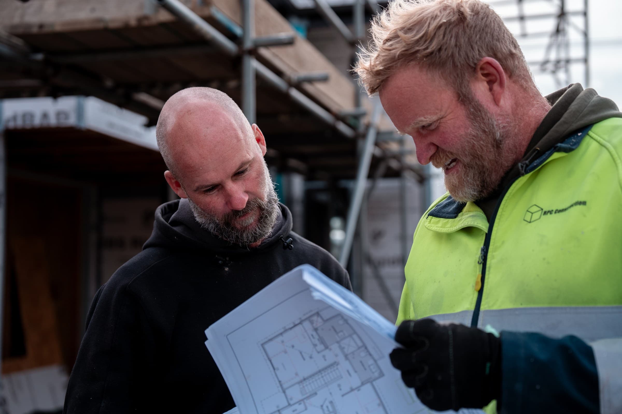 Two people looking at an open architectural floor plan held between them on a building site, one in an RPC Construction hi-vis jacket and work gloves, scaffolding and building wrap behind them