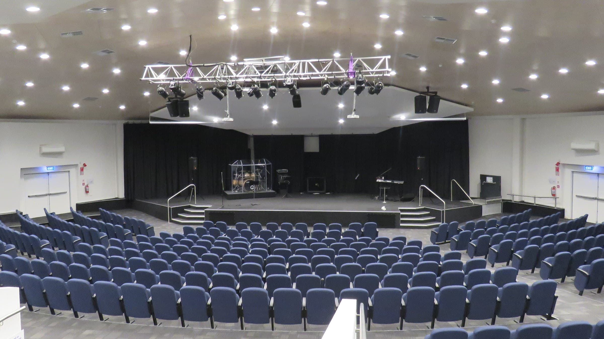 A large auditorium with banked rows of blue upholstered seats facing a dark stage, a lighting truss suspended above and a drum kit and keyboard set up under the house lights