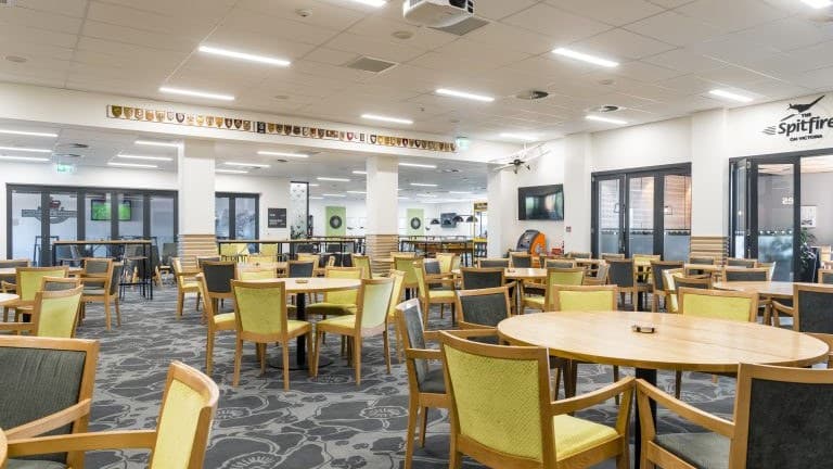 A club dining room with round timber tables and green and grey upholstered chairs on patterned carpet, honour boards along one wall and a frieze of squadron badges near the ceiling