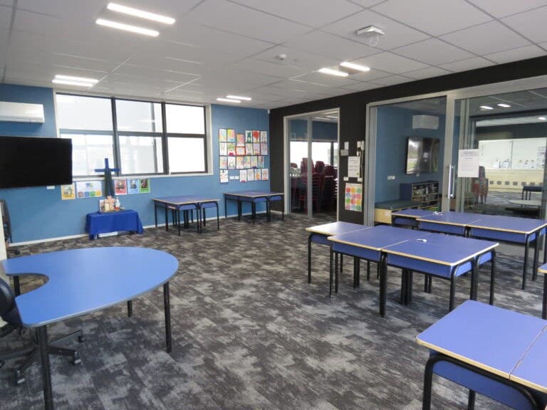 A school classroom with blue-topped tables and stacking chairs set out across patterned grey carpet, a wall-mounted screen and children's artwork pinned along a blue wall, and a glazed partition opening into a second teaching space beyond