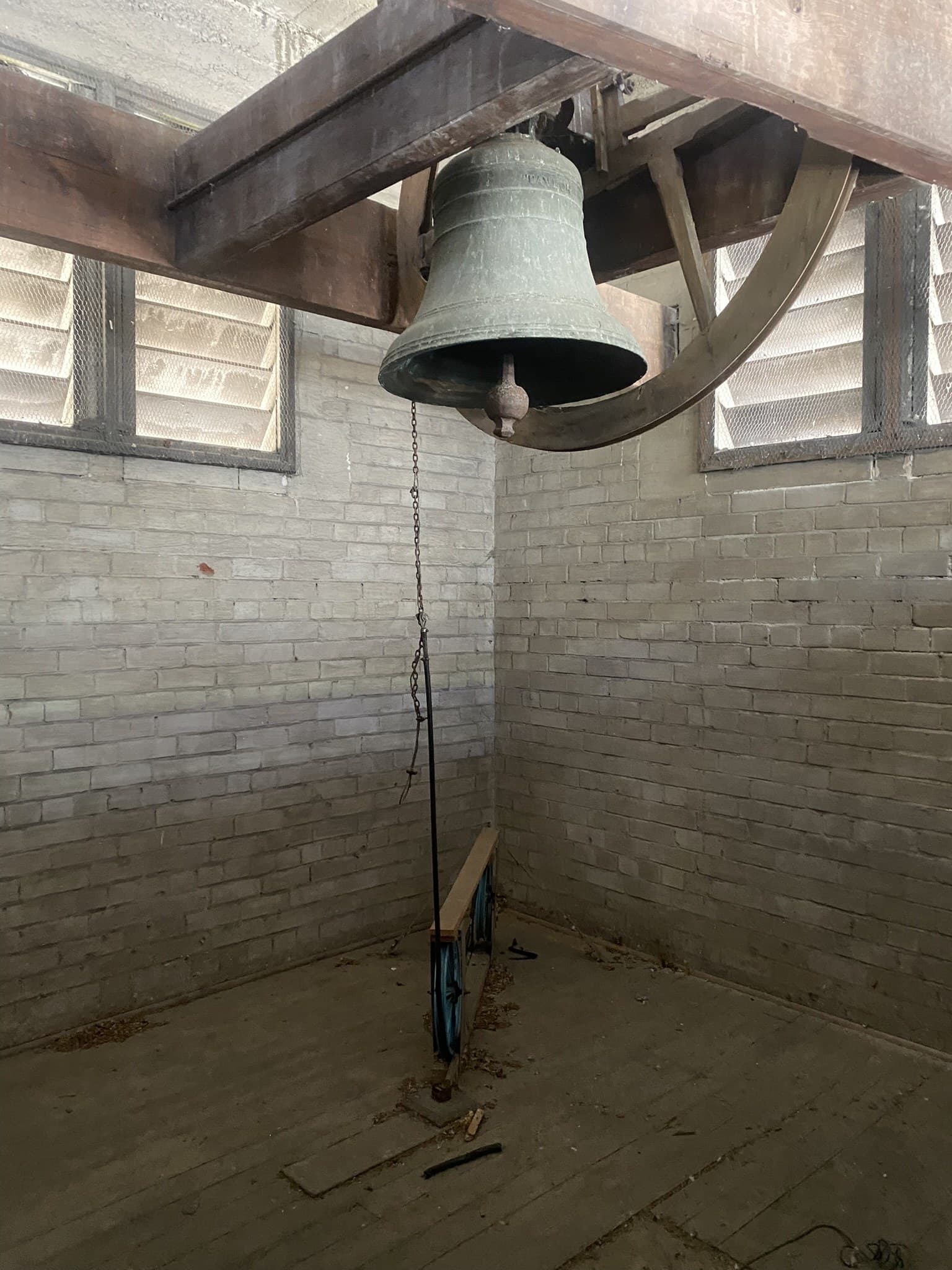 A large bronze bell hanging in a timber headstock and wheel inside a whitewashed brick bell tower, with a rope dropping to the bare boarded floor below and louvred openings either side
