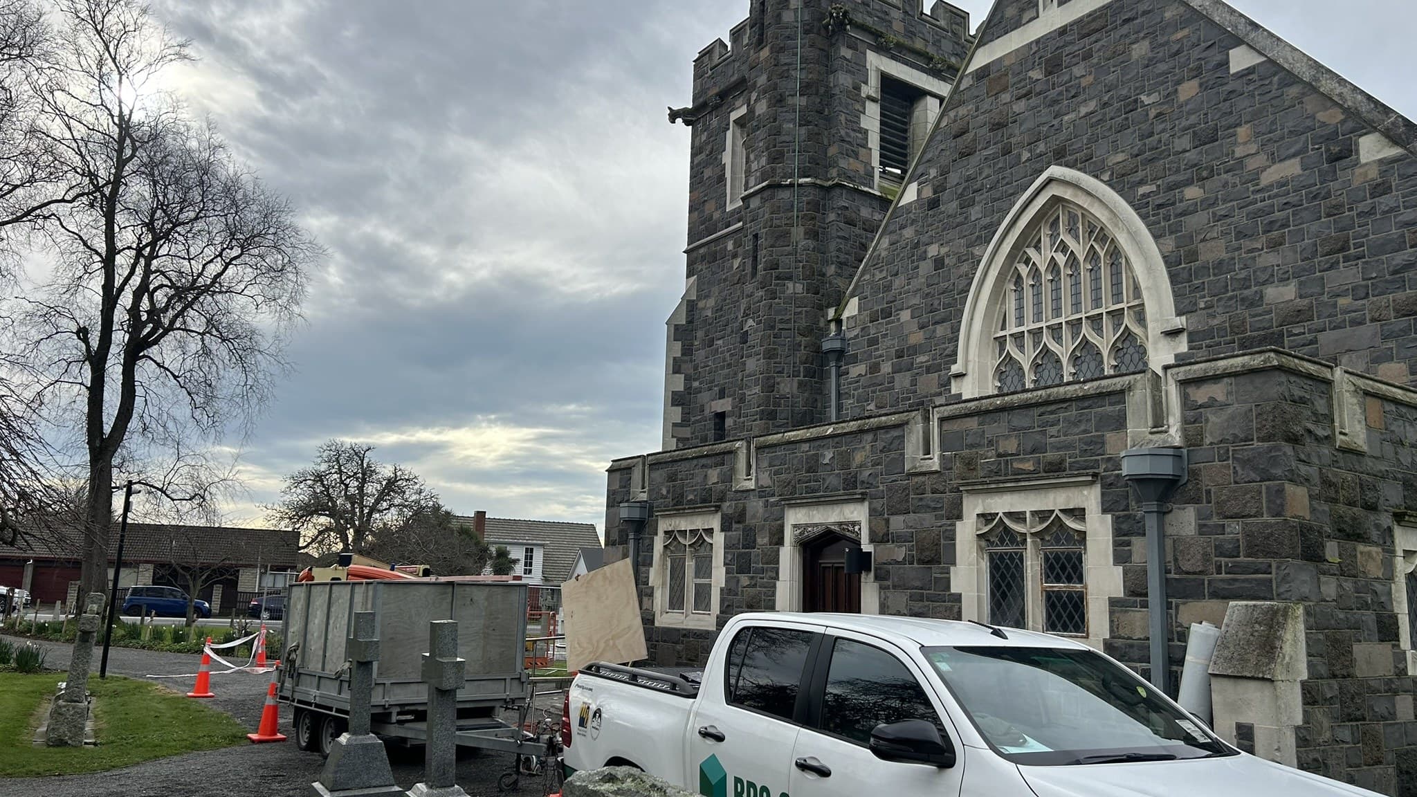A stone Gothic church with a crenellated tower, an RPC Construction ute and a loaded flat-deck trailer parked on the drive beside it, traffic cones and a bunting line across the drive, headstones in the foreground