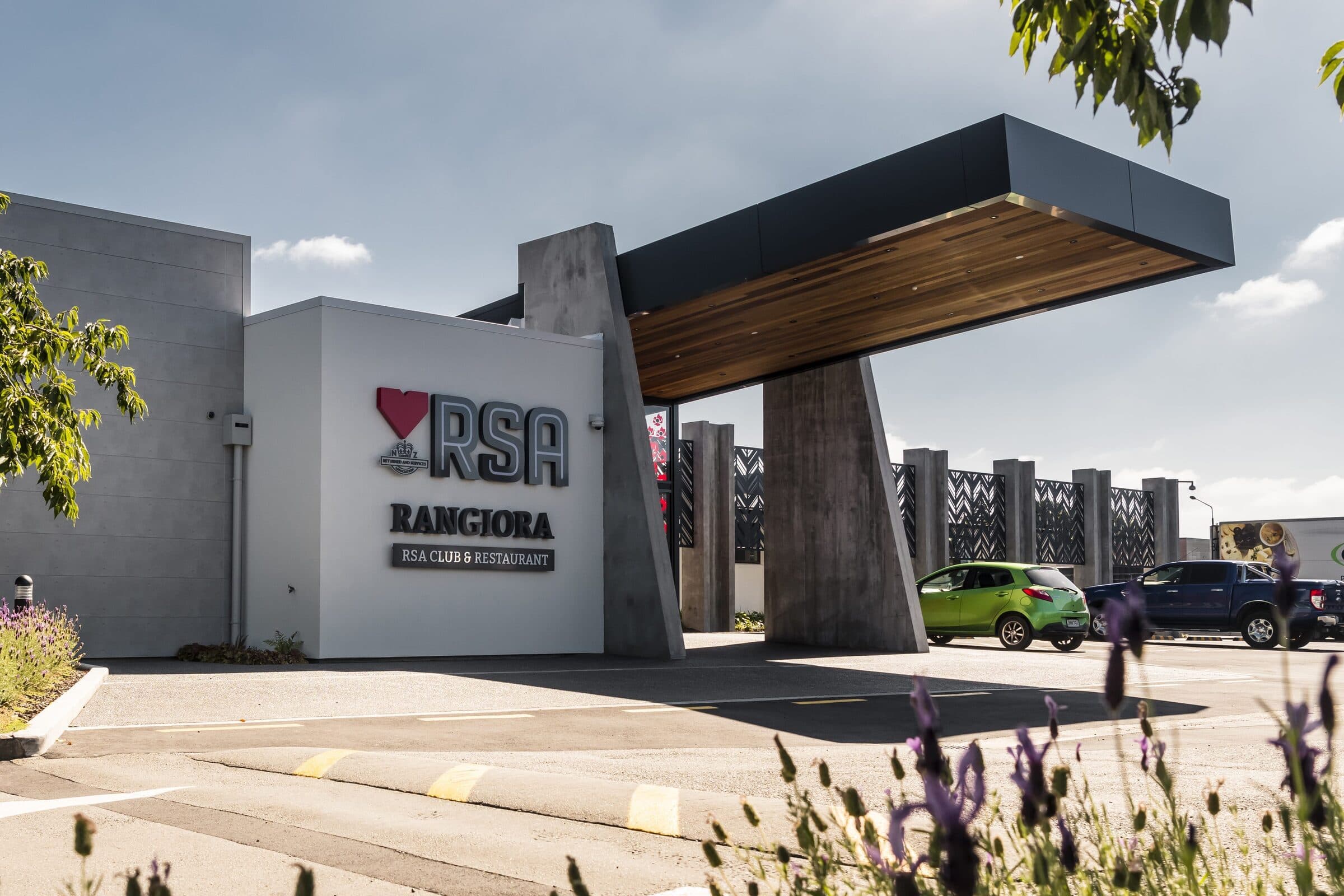 The completed entrance to the Rangiora RSA Club and Restaurant in bright sun: raised lettering on a white rendered wall, a timber-lined canopy carried on angled concrete columns over the entry, parked cars beyond and lavender in flower along the kerb