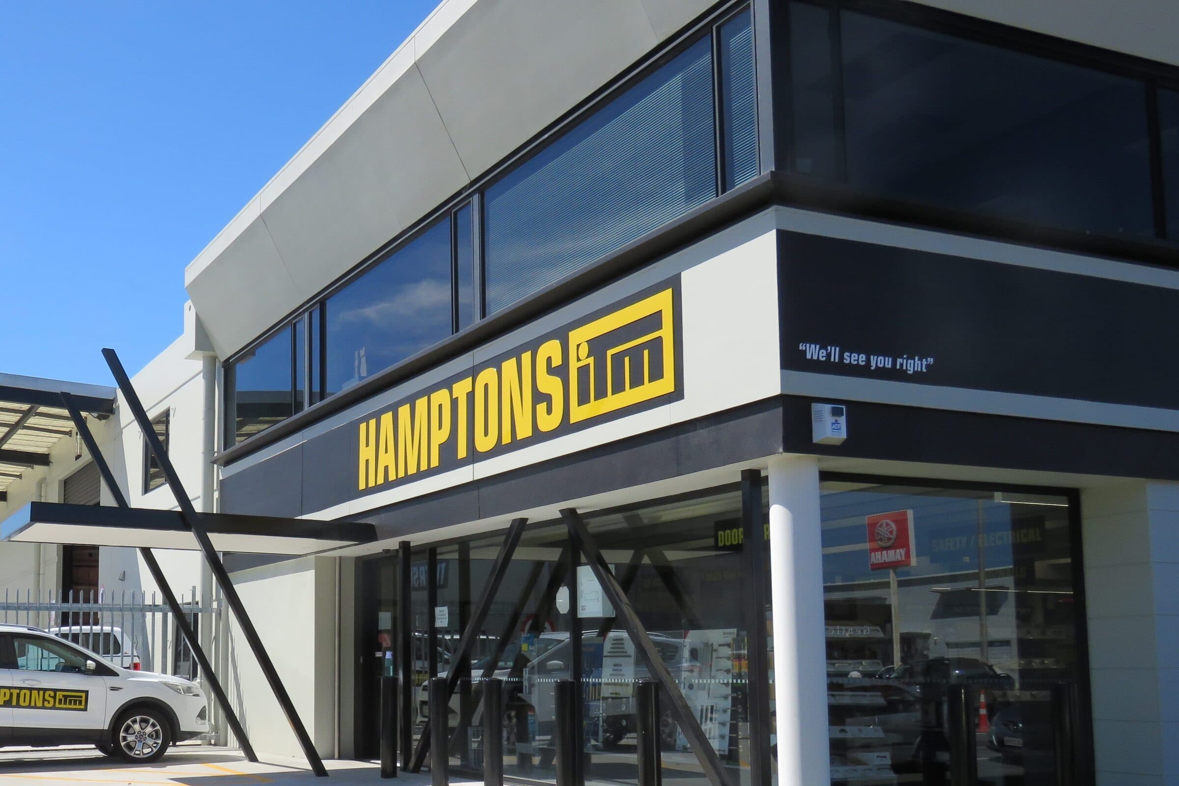 The completed Hamptons ITM store frontage in bright sun: yellow lettering on a dark fascia above full-height glazing, angled black steel bracing across the front of the building, stock shelving visible inside and a branded vehicle parked at the kerb