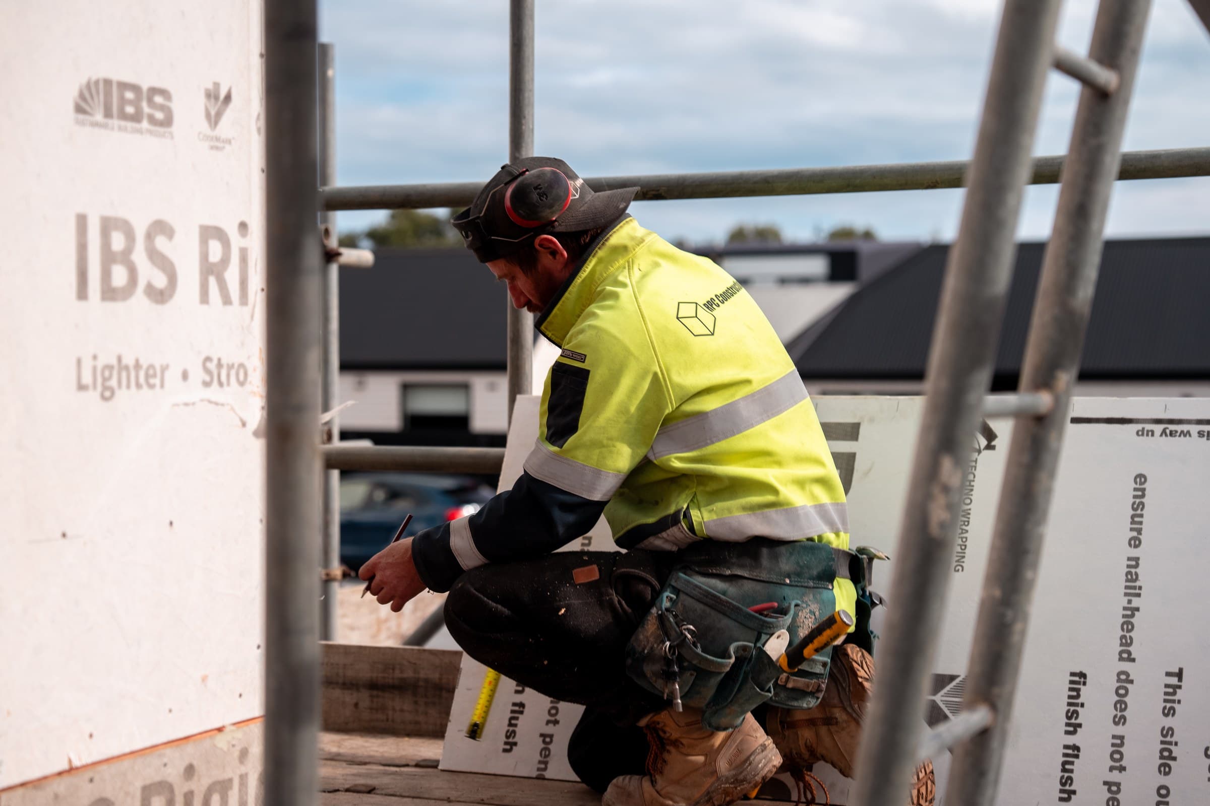 A worker in an RPC Construction hi-vis top and tool belt crouching on a scaffold platform on a building under construction, marking a sheet of building board with a pencil, a tape measure on the deck beside him and scaffold tubes across the frame