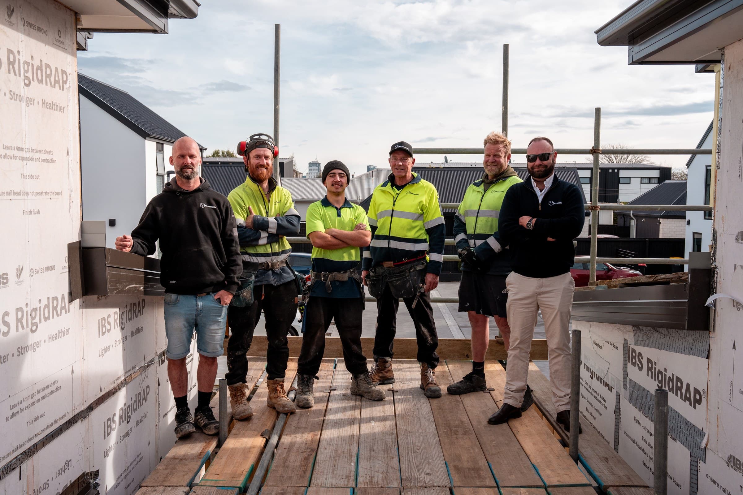 Six people standing together on the timber deck of a house under construction, four in high-visibility workwear and boots, one in a dark jumper and one in business clothes, with building wrap on the walls behind them