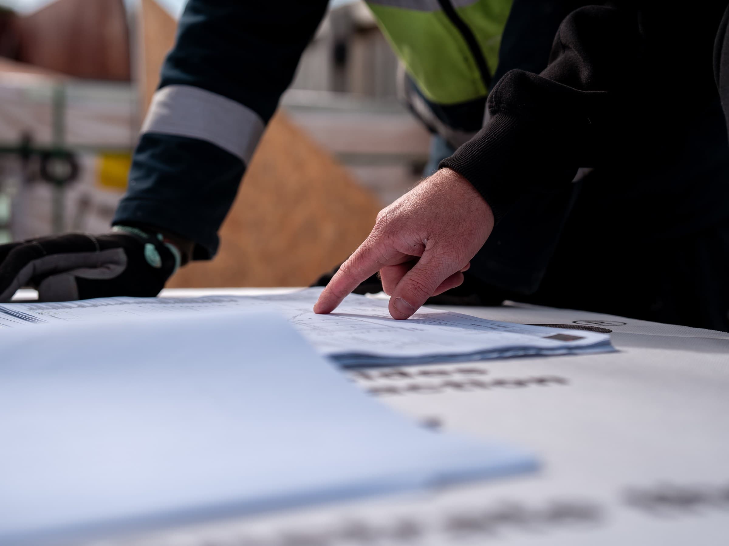 Close on two people's hands over a set of drawings laid out on a sheet of building board on a site, one bare hand pointing at the sheet, the other in a work glove