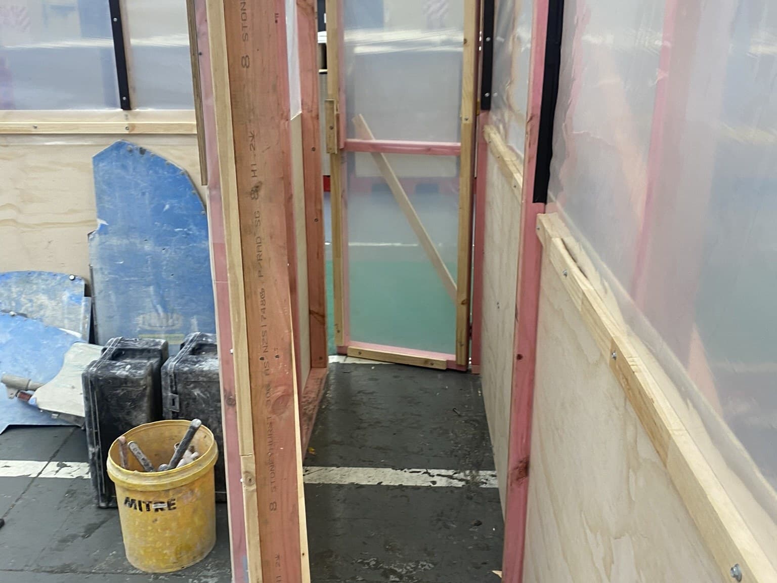 The inside of a partly built room with exposed timber framing, polythene sheeting over the openings and plywood lining fixed to the lower walls, a bucket and tool cases on the floor