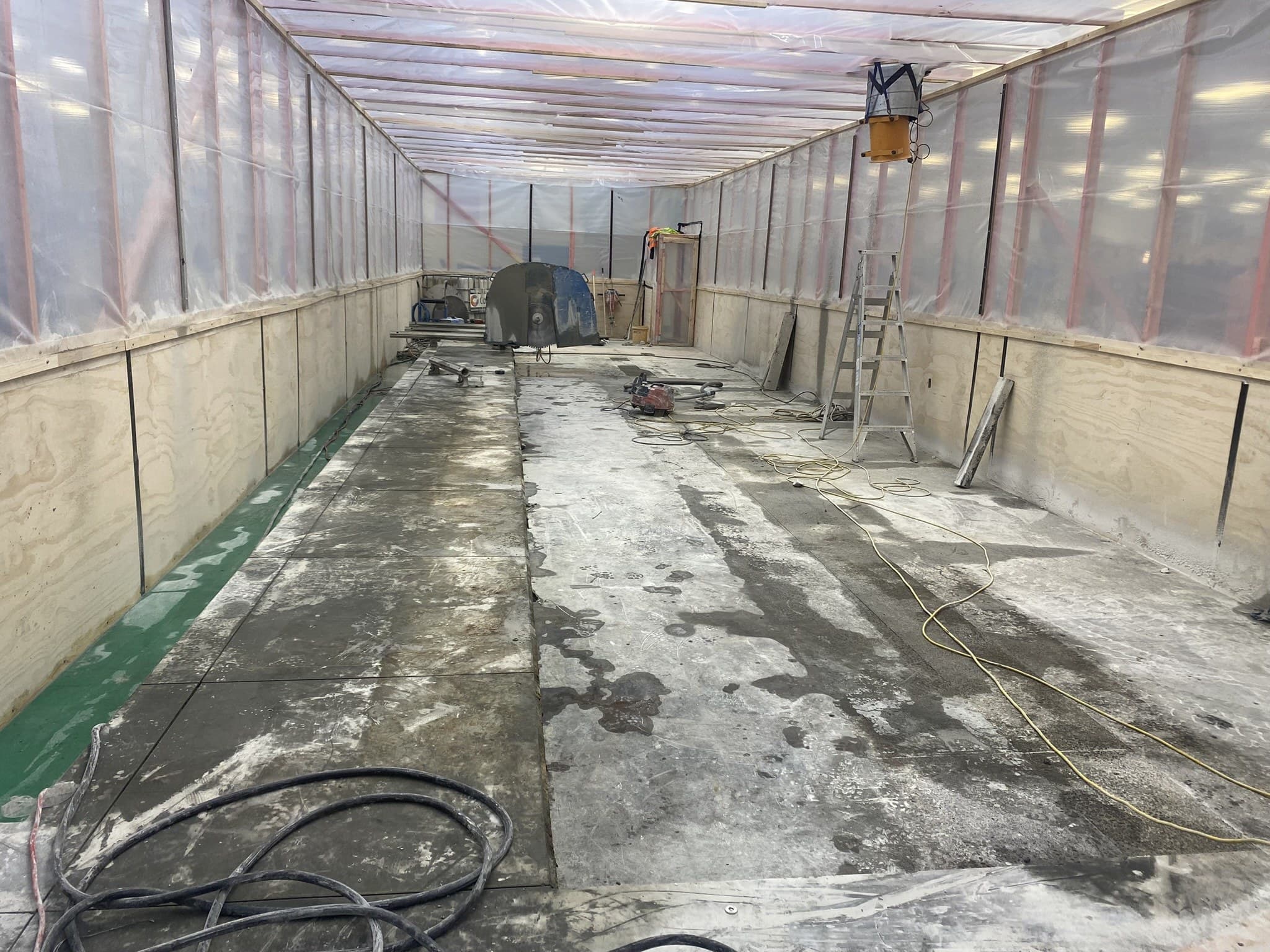 A work area sheeted floor to ceiling in plywood and plastic containment, its concrete floor ground back to bare slab, with a large floor saw and spare blades at the far end, a grinder and vacuum hose set down on the slab, a stepladder standing clear, and leads and hoses run out across the floor