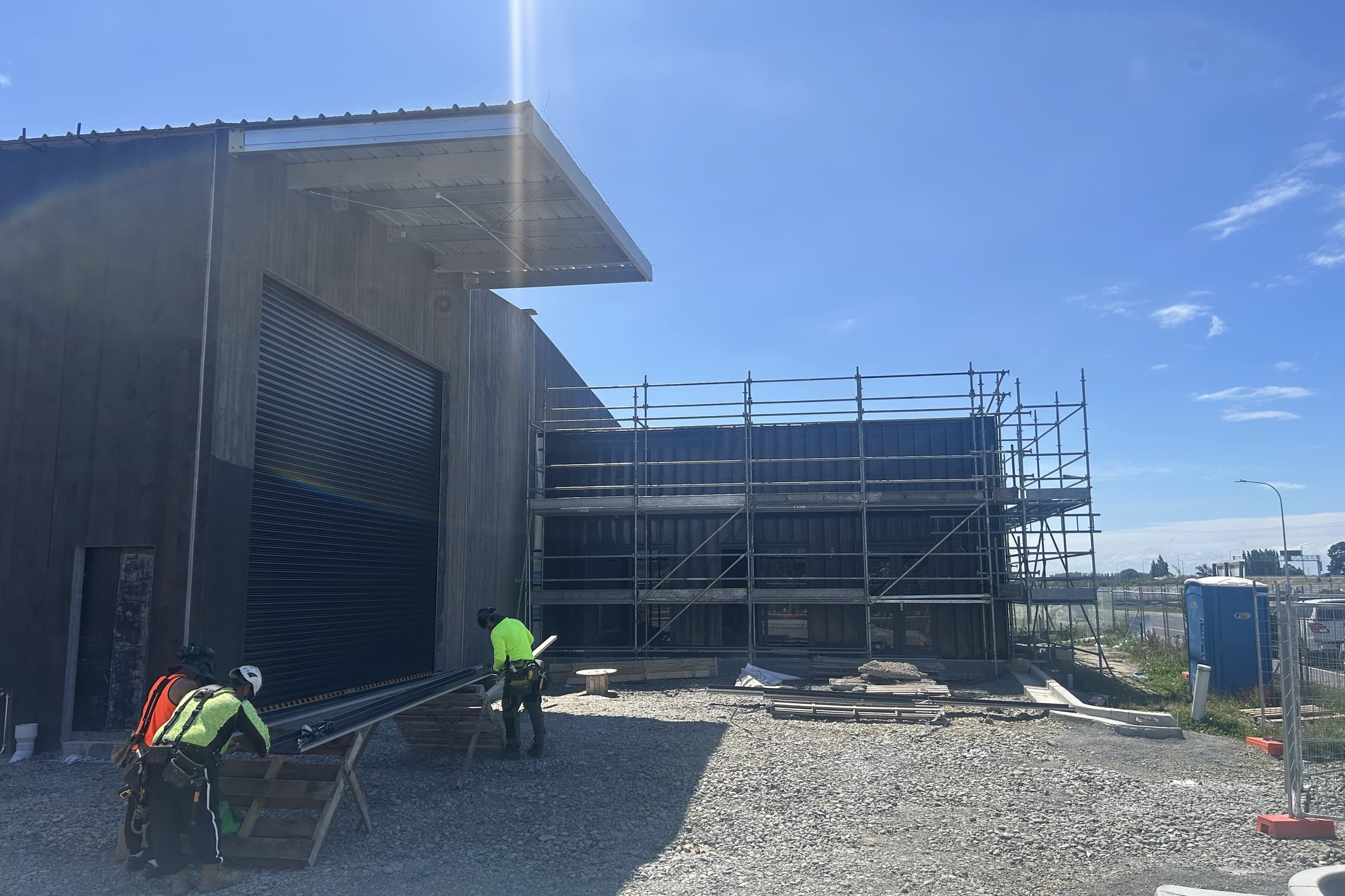 Two workers in high-visibility clothing cutting material on trestles outside a part-clad building, with scaffolding erected across the two-storey section beside them
