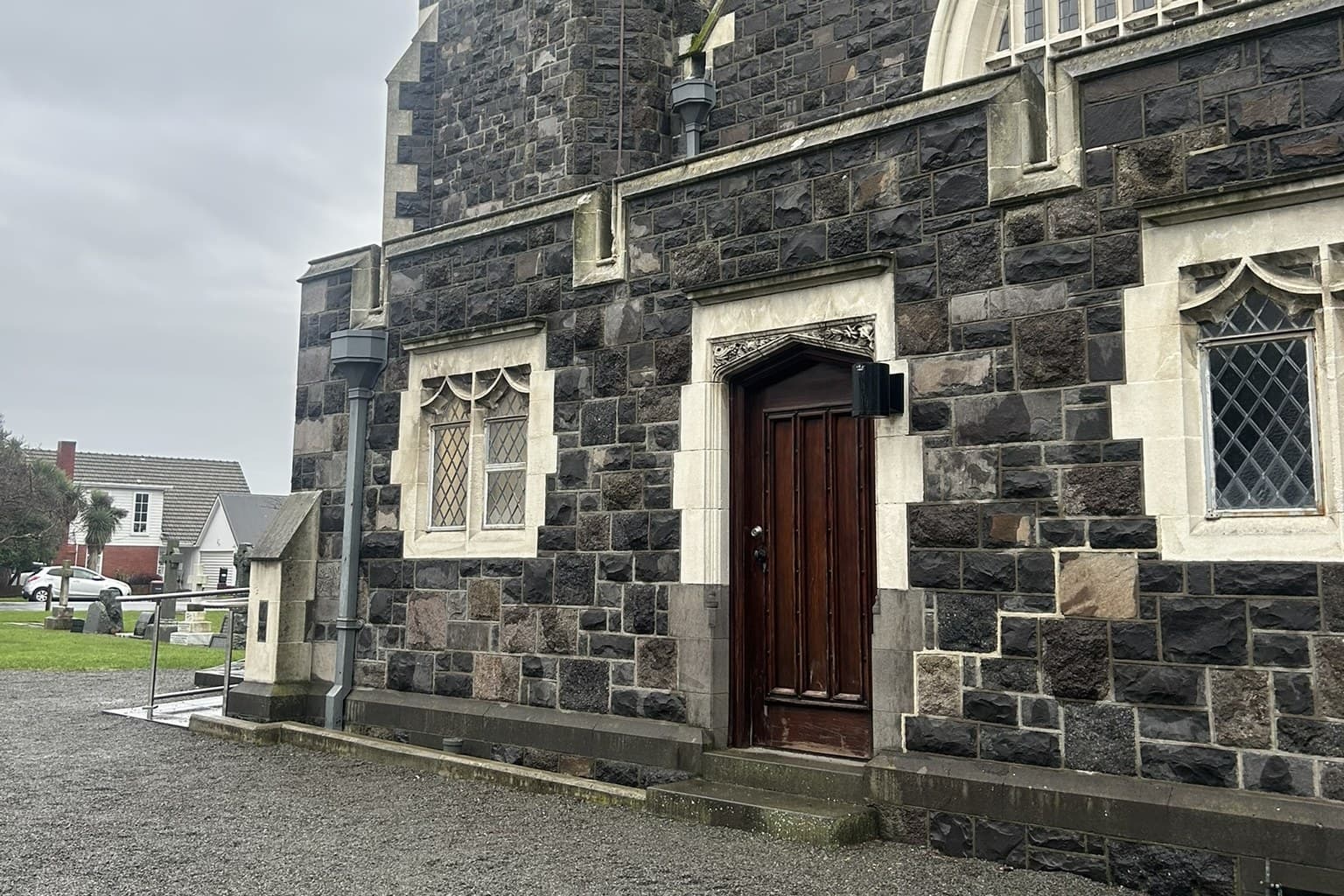 The side of a stone church seen along a gravel path, with a closed timber door in a carved stone surround, a handrail at the step up to it, leaded windows either side, and headstones on the grass and houses across the road beyond