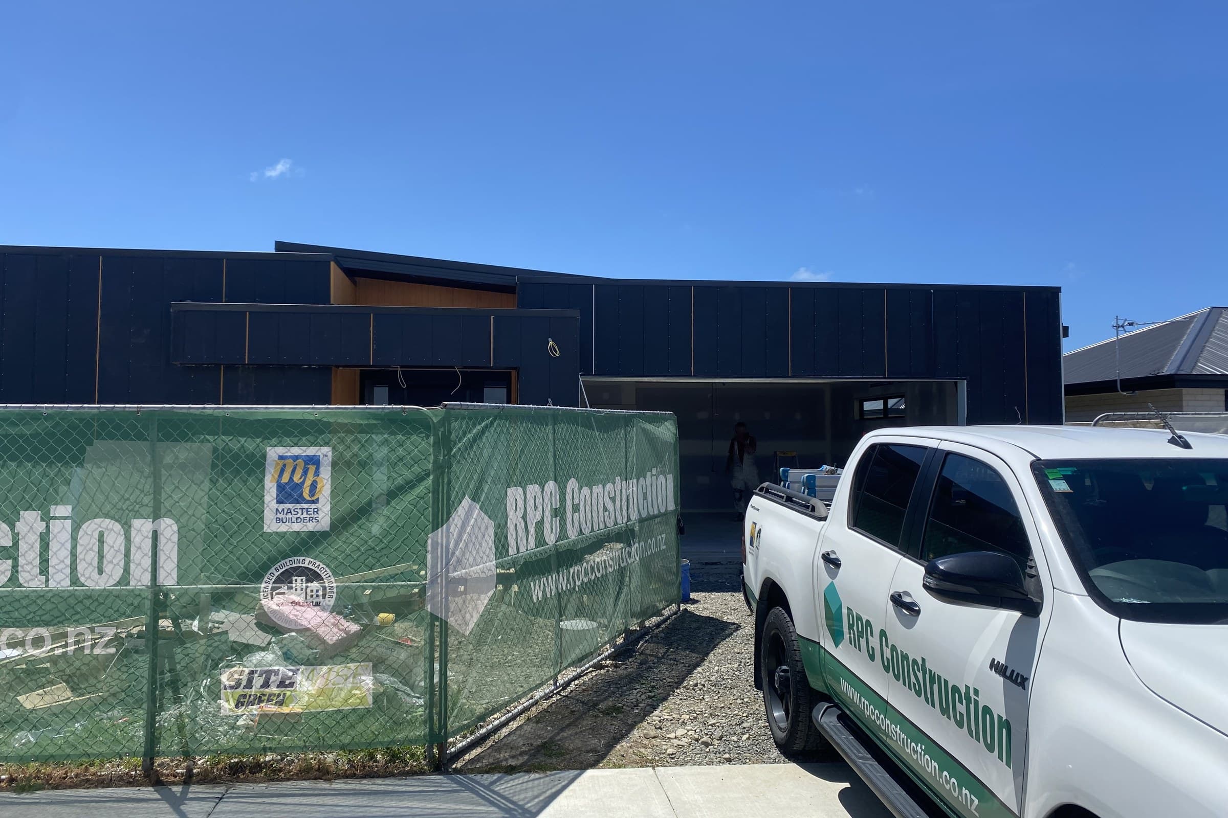 Temporary site fencing with green RPC Construction scrim stretched across it, a white RPC ute parked alongside, and a dark-clad building under construction behind the gates