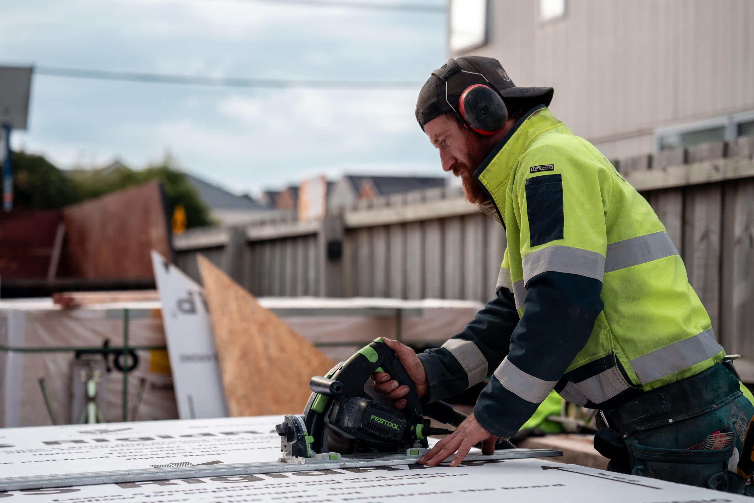A carpenter in a high-visibility jacket and ear defenders running a circular saw along a guide rail across a sheet of insulated panel, with a timber site boundary fence behind