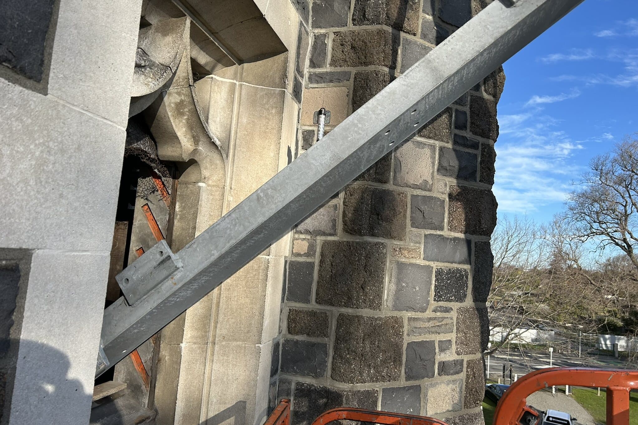 A new galvanised steel channel being manoeuvred against the basalt and limestone wall of a church tower, beside a stone-traceried opening with its timber louvre partly removed